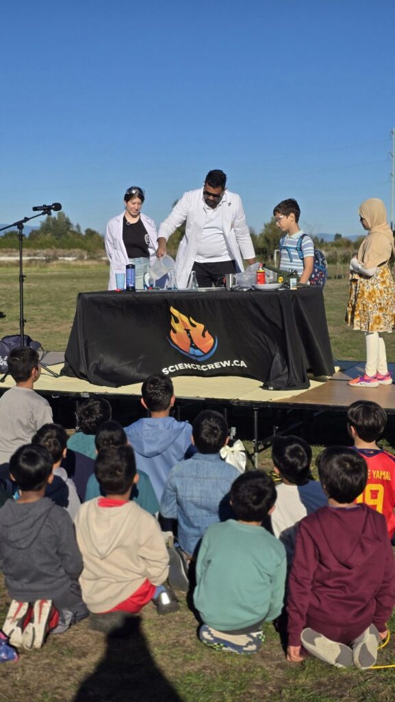 Children seated on the grass watching a science show on the outdoor stage at the AZIA Family BBQ